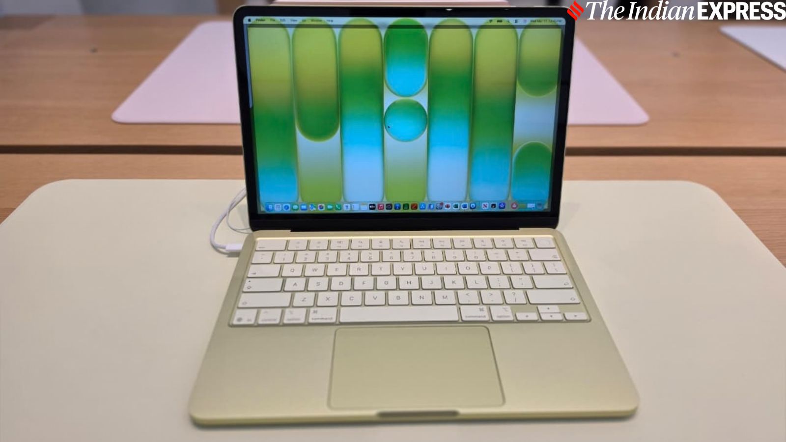 Gold-colored laptop on a white desk, open to a bright blue‑green wallpaper with The Indian Express watermark in the corner.