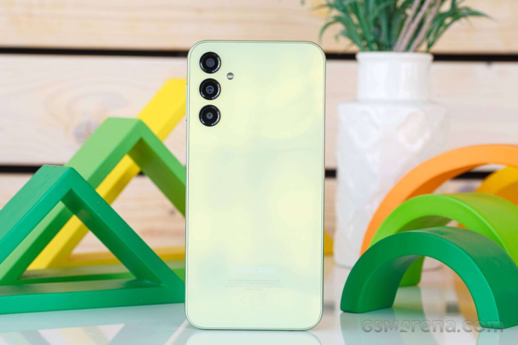 Pale green Samsung smartphone standing upright on a reflective surface, showing its triple-camera module with a white planter and colorful green arches in the background.