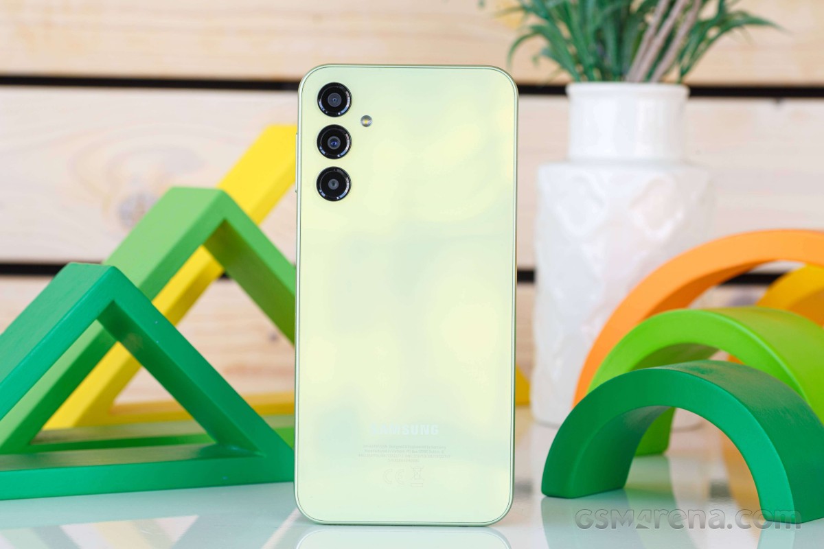 Pale green Samsung smartphone standing upright on a reflective surface, showing its triple-camera module with a white planter and colorful green arches in the background.