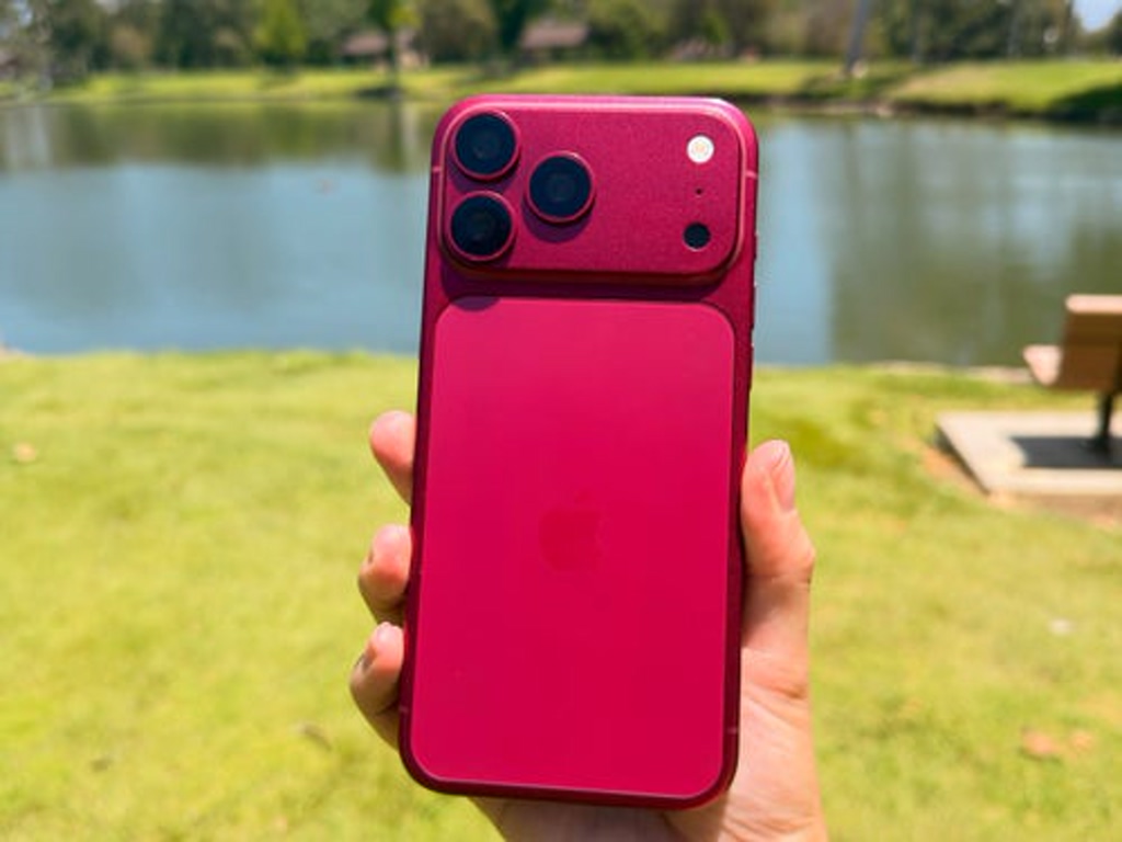Person holding a pink iPhone with triple camera setup outdoors near a lake in a grassy park setting