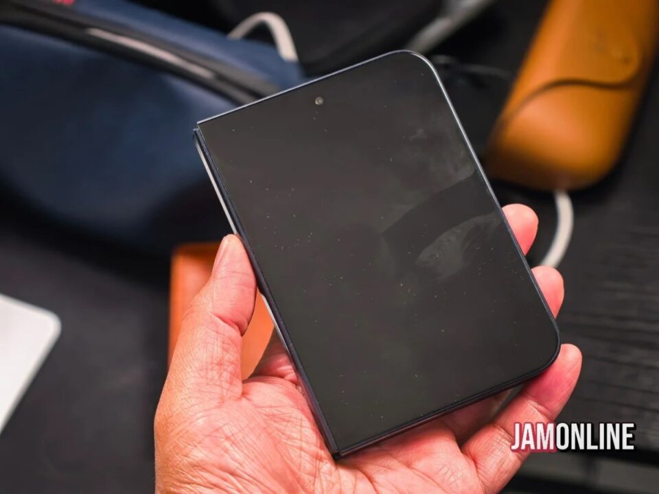 Close-up of a hand holding a black smartphone with a glossy, blank screen and a blurred bag in the background, JamOnline watermark visible.