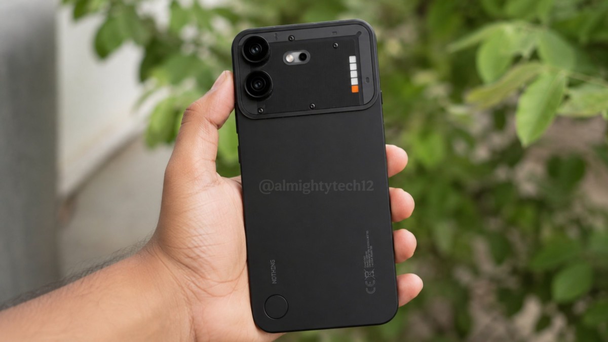 Back of a black smartphone showing a raised triple-camera module held in a hand outdoors against a green leafy background, watermark visible on the back