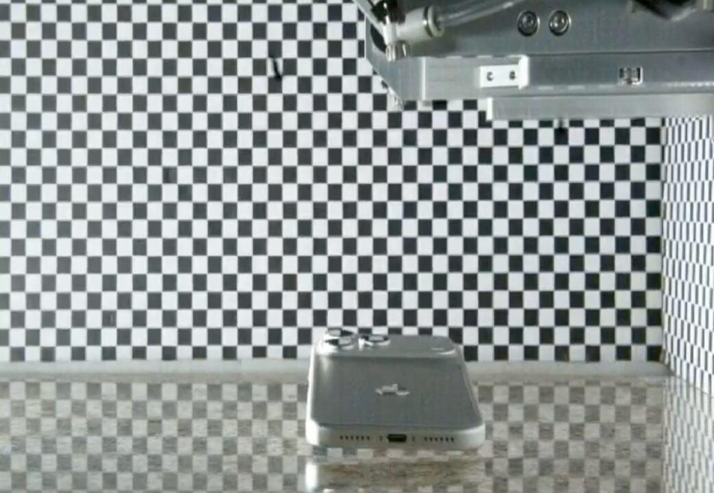 Silver iPhone resting on a reflective table with a black-and-white checkerboard backdrop, suggesting a testing or calibration setup.
