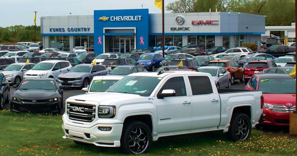 White GMC Sierra pickup parked prominently in front of a busy Chevrolet-Buick-GMC dealership showroom with a crowded car lot behind it.