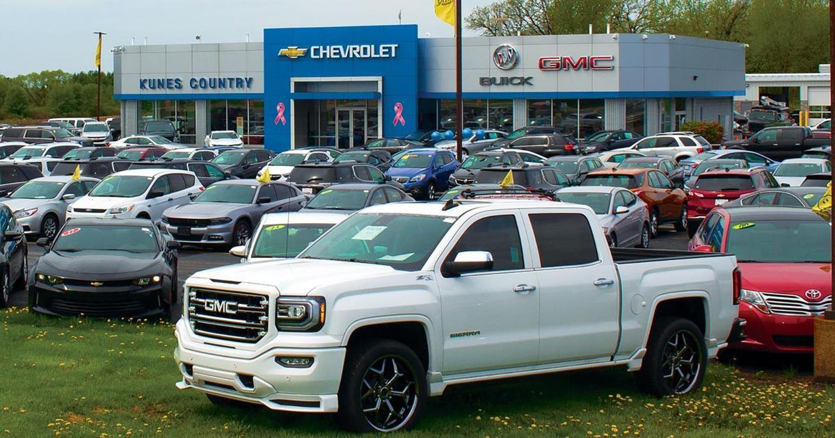 White GMC Sierra pickup parked prominently in front of a busy Chevrolet-Buick-GMC dealership showroom with a crowded car lot behind it.
