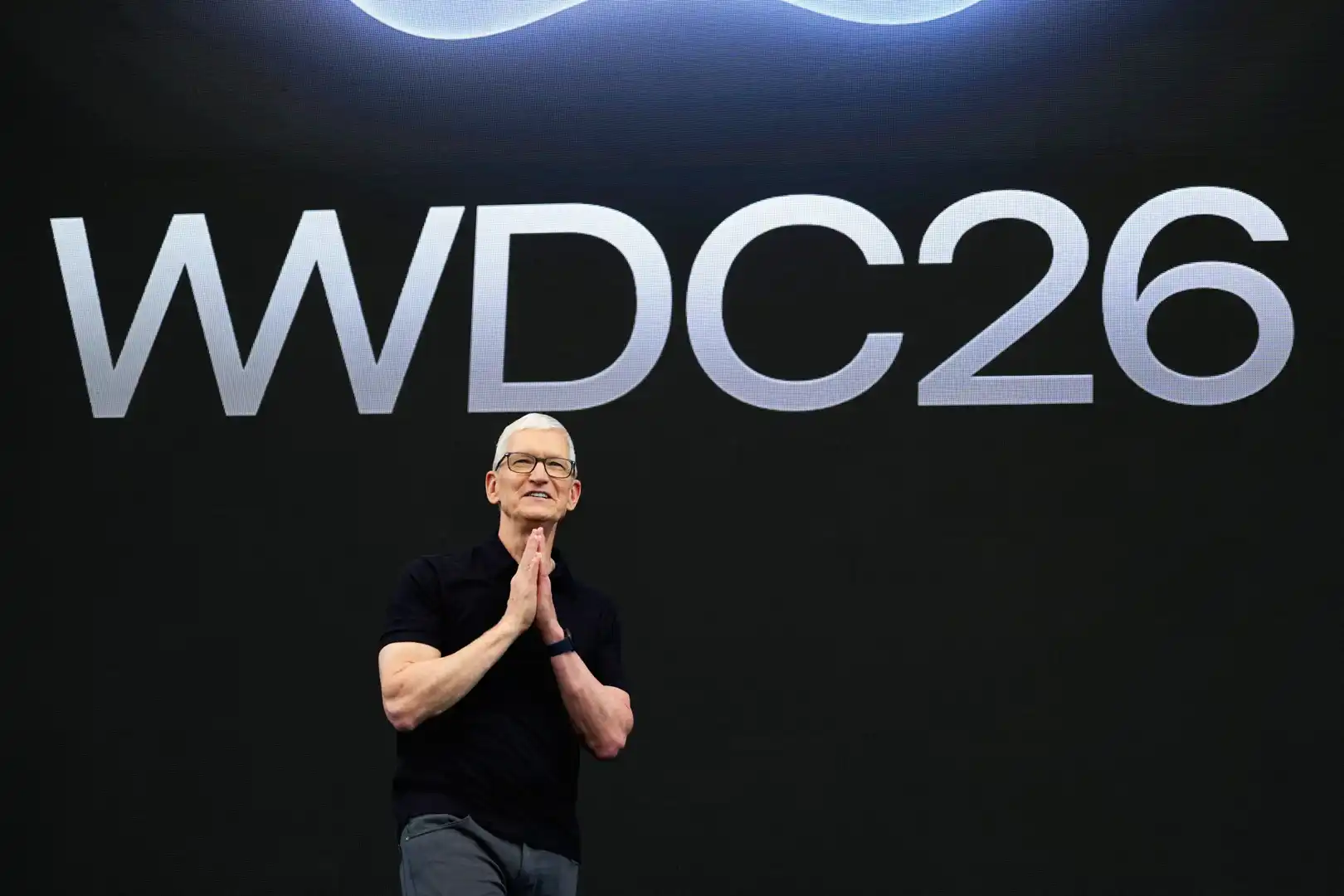 Speaker on a dark stage with a large WWDC26 sign behind him, clasping hands in front.