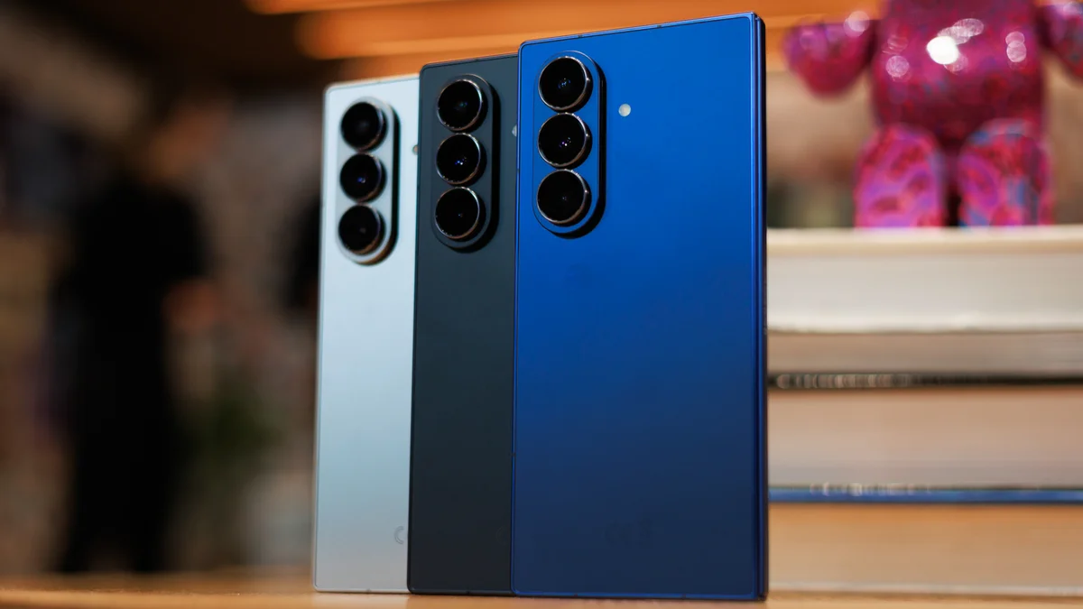 Three smartphones standing upright on a wooden surface: blue, dark gray, and silver backs with prominent triple-camera modules.