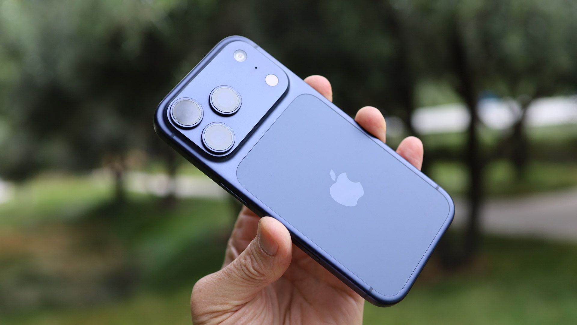 Blue iPhone held in a hand outdoors, showing the triple-camera array on the back.