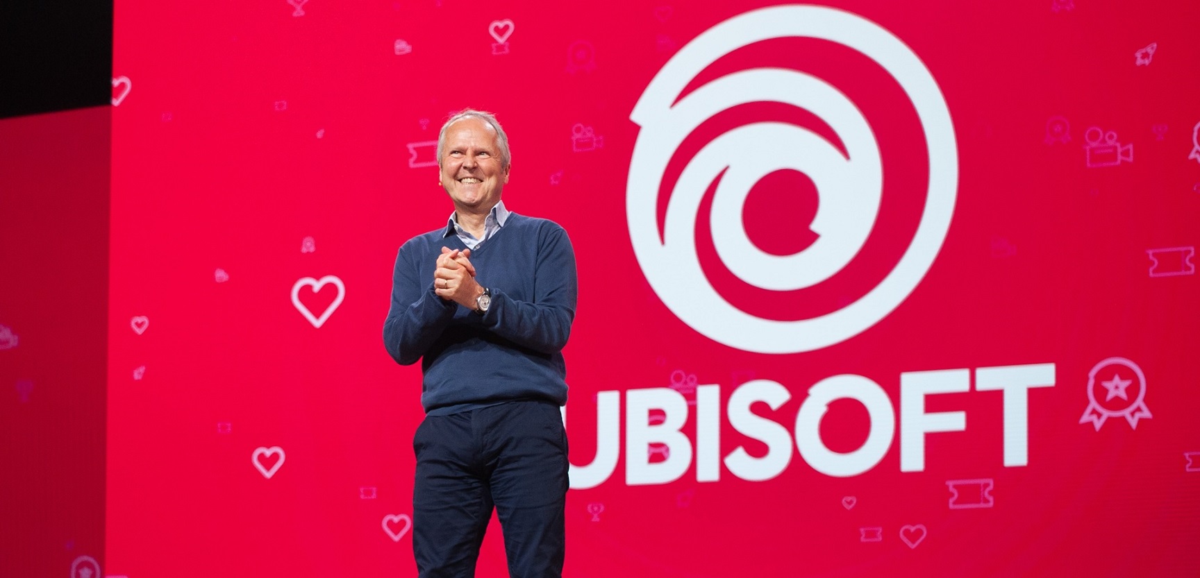 Smiling Man in a Blue Sweater Stands on a Stage in Front of a Large White Ubisoft Logo on a Red Backdrop