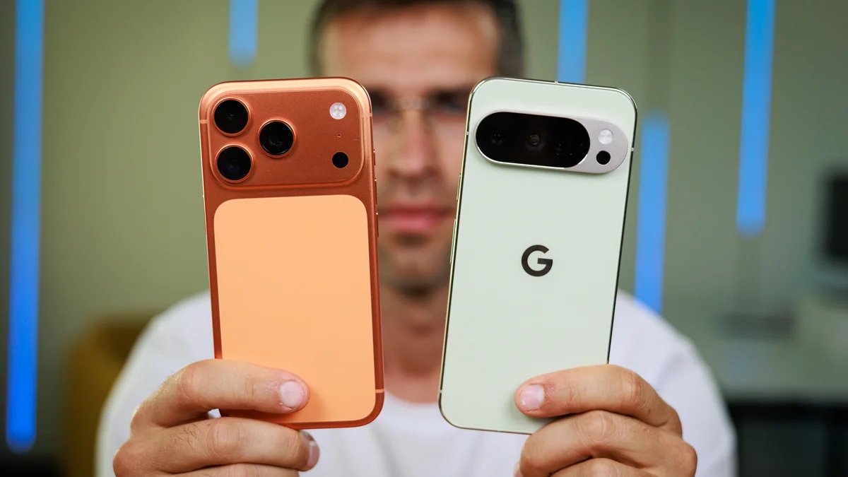 Person holding two smartphones with large rear cameras, orange and light green backs, in a tech setting.