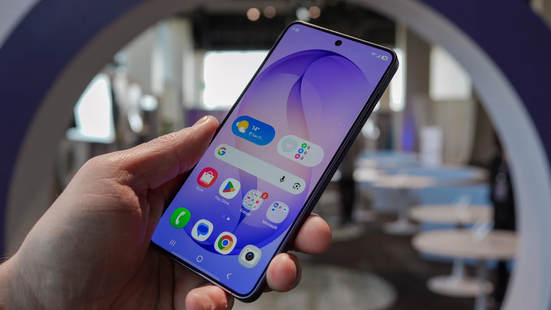 Hand holding a smartphone with a purple wallpaper and visible app icons, blurred indoor background beyond the screen.
