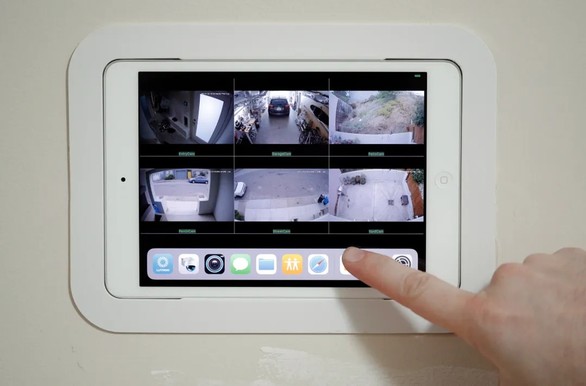 Wall-mounted tablet displaying a multi-camera security feed grid; a finger taps the app dock at the bottom panel.