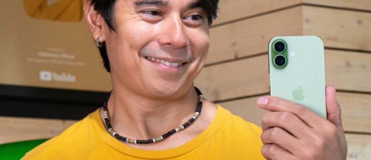 Smiling man in a yellow shirt holding a mint-green iPhone and taking a selfie, with a wooden wall in the background.