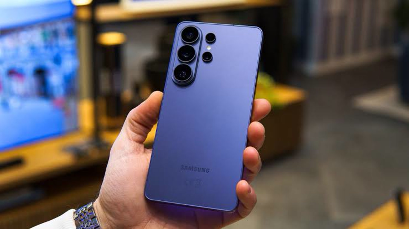 Blue Samsung smartphone held in a person's hand, showing a quad-camera module on the back in a blurred indoor setting.