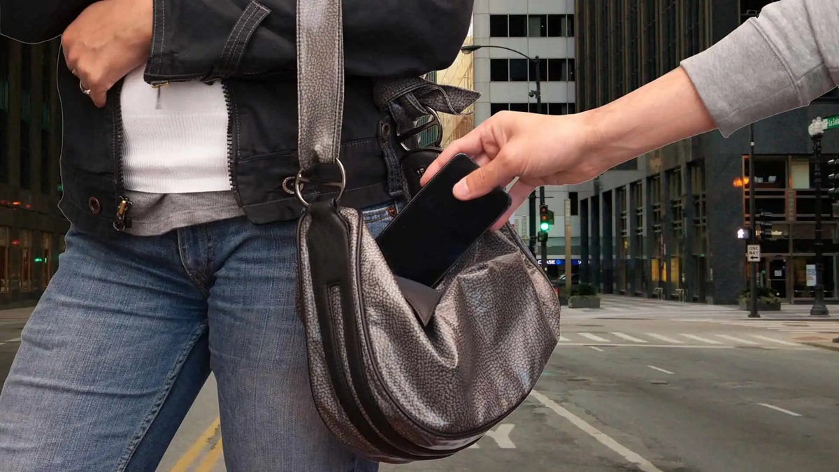 A hand reaches into a metallic gray purse to grab a smartphone on a city street backdrop.