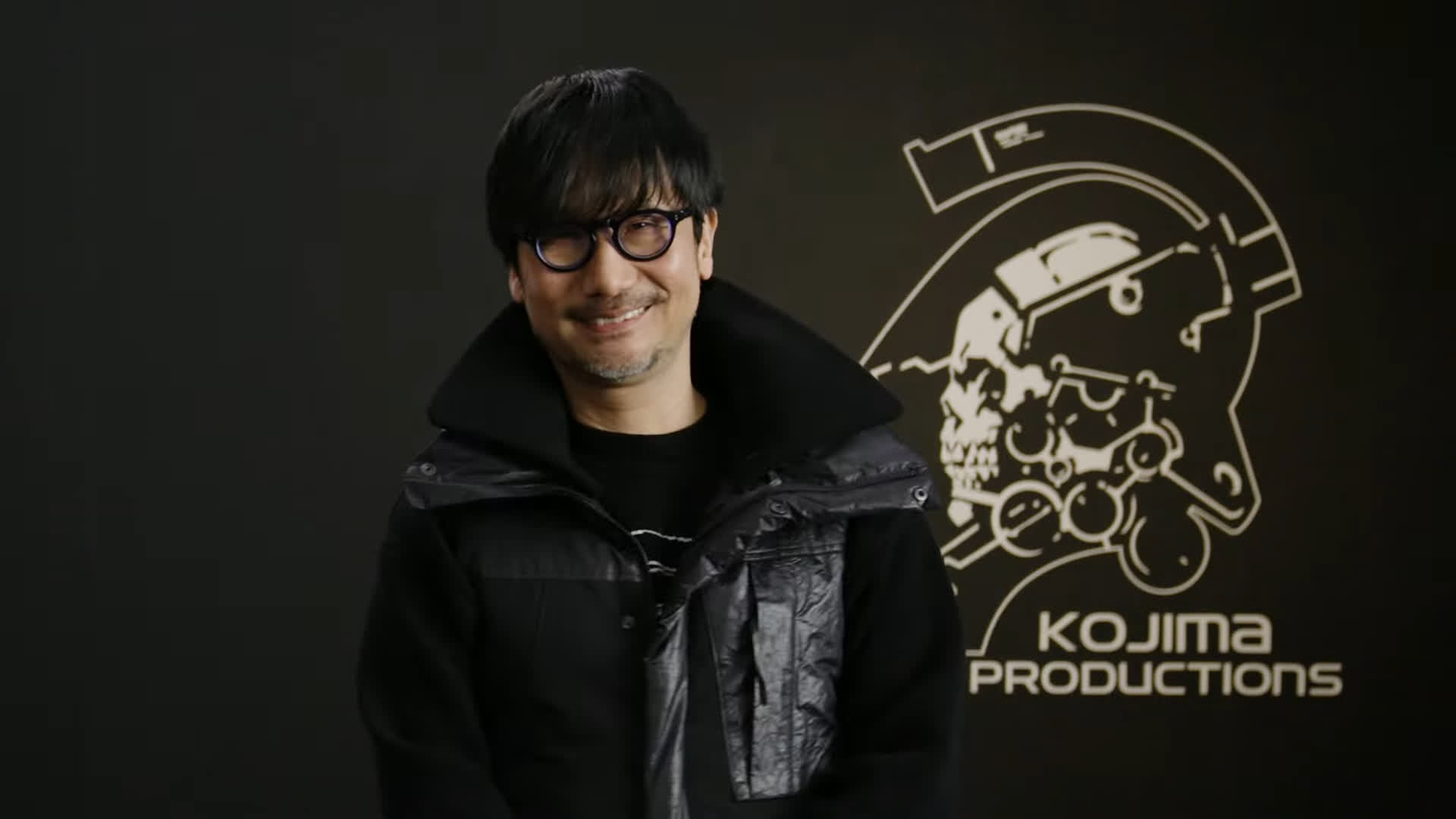 Smiling Man Wearing Round Glasses and a Black Jacket Stands in Front of a Kojima Productions Backdrop