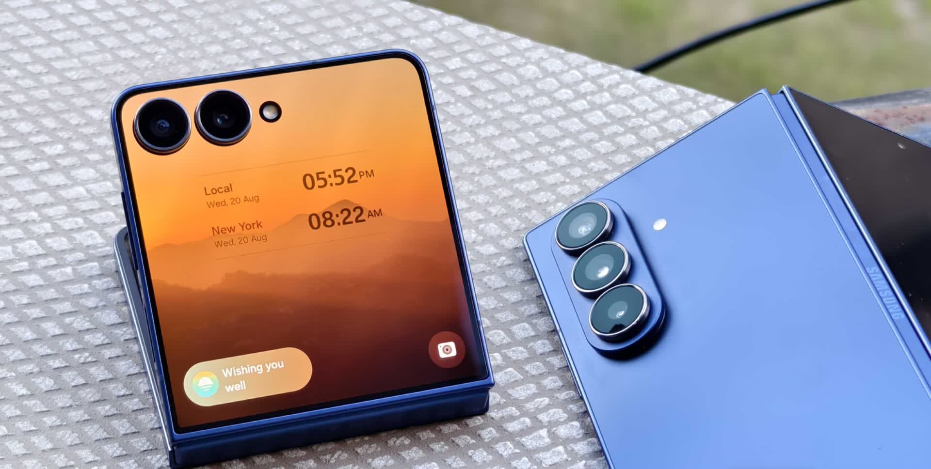 Blue foldable smartphone on a textured surface, front screen showing sunset wallpaper, time and weather widgets; triple-camera module visible on the back edge.