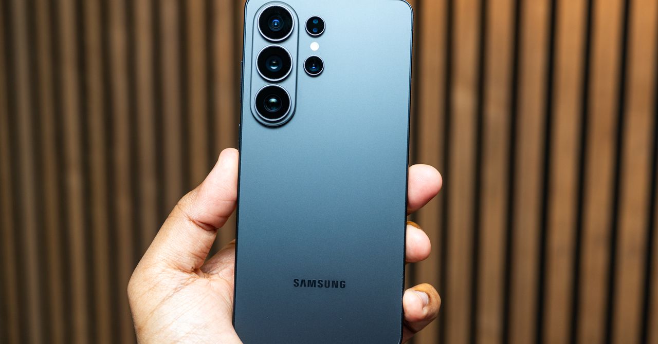 Back of a blue Samsung Galaxy phone with a large multi‑lens camera module being held by a hand, wooden striped background visible.