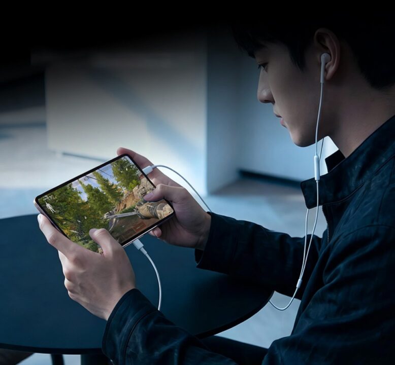 Person wearing white earbuds and a black jacket plays a forest-themed game on a smartphone at a table, focused on the screen.
