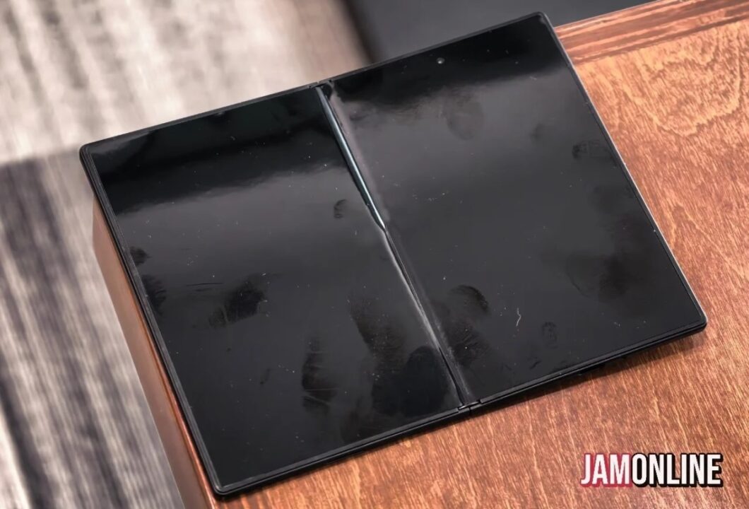 Open foldable device with two glossy black screens laid flat on a wooden desk; JAMONLINE watermark visible on corner.
