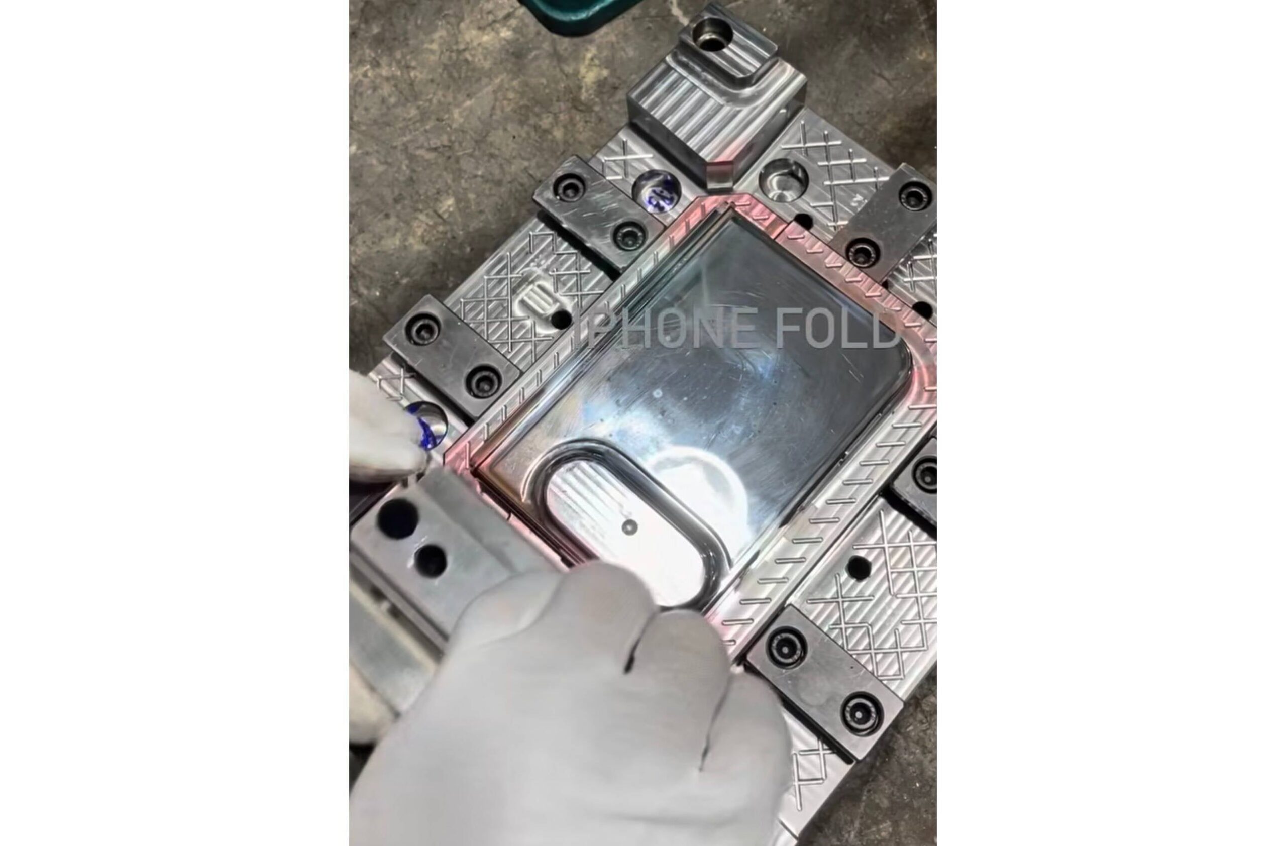 1000049414 Close-up of a metal die or mold with a diamond-pattern surface and clamps, a gloved hand in the foreground; watermark text 'PHONE FOLD' visible.