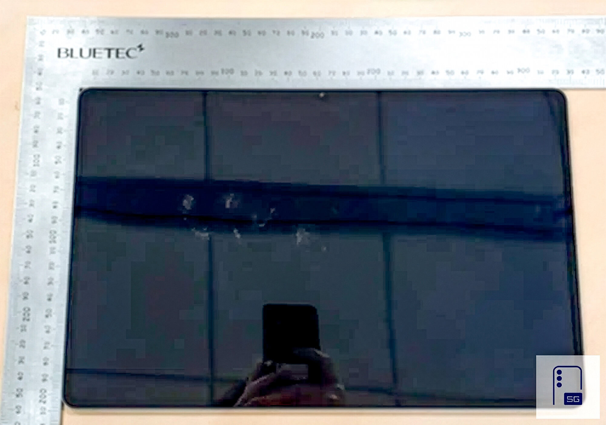 Top-down view of a dark smartphone on a light desk, with metal rulers along the top and left edges and a 5G sticker in the bottom-right corner.