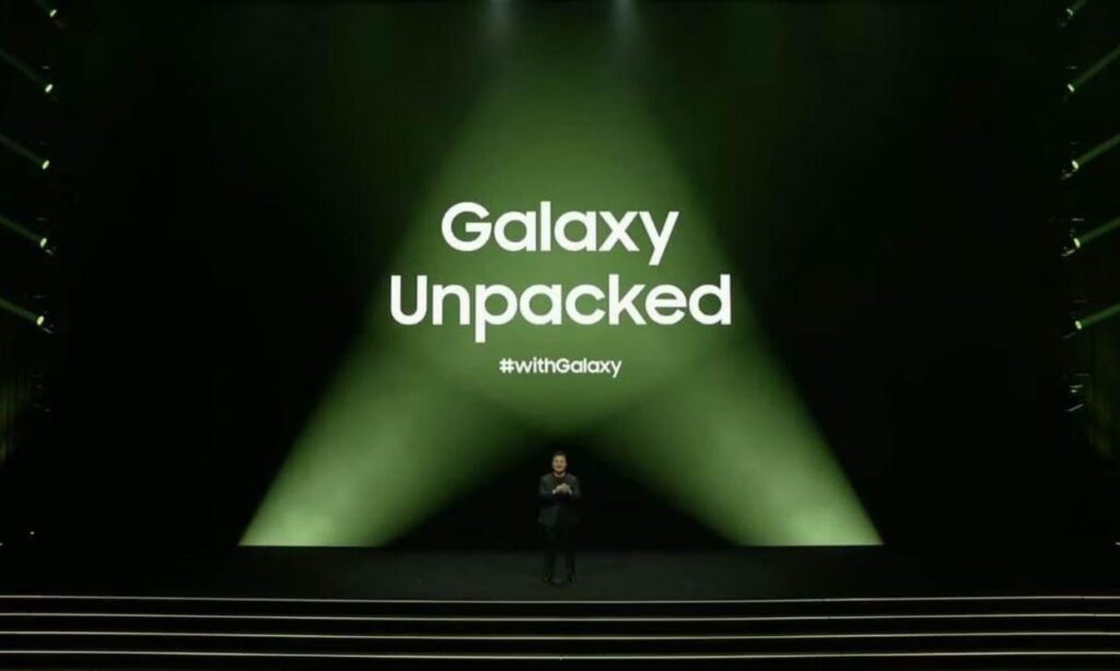 Presenter on a dark stage with a large green spotlight backdrop reading 'Galaxy Unpacked' and the hashtag '#withGalaxy' nearby, at an event.