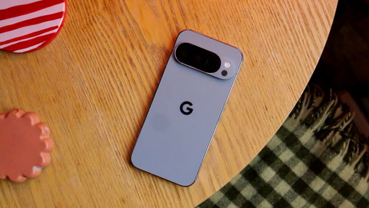 Blue Google Pixel phone with a dual‑camera bar lies on a wooden table; a red striped cup and green plaid blanket are nearby.