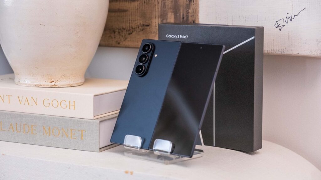 Samsung Galaxy Z Fold7 in dark blue on a clear display stand beside its black box, with decorative books and a vase in the background (product showcase).