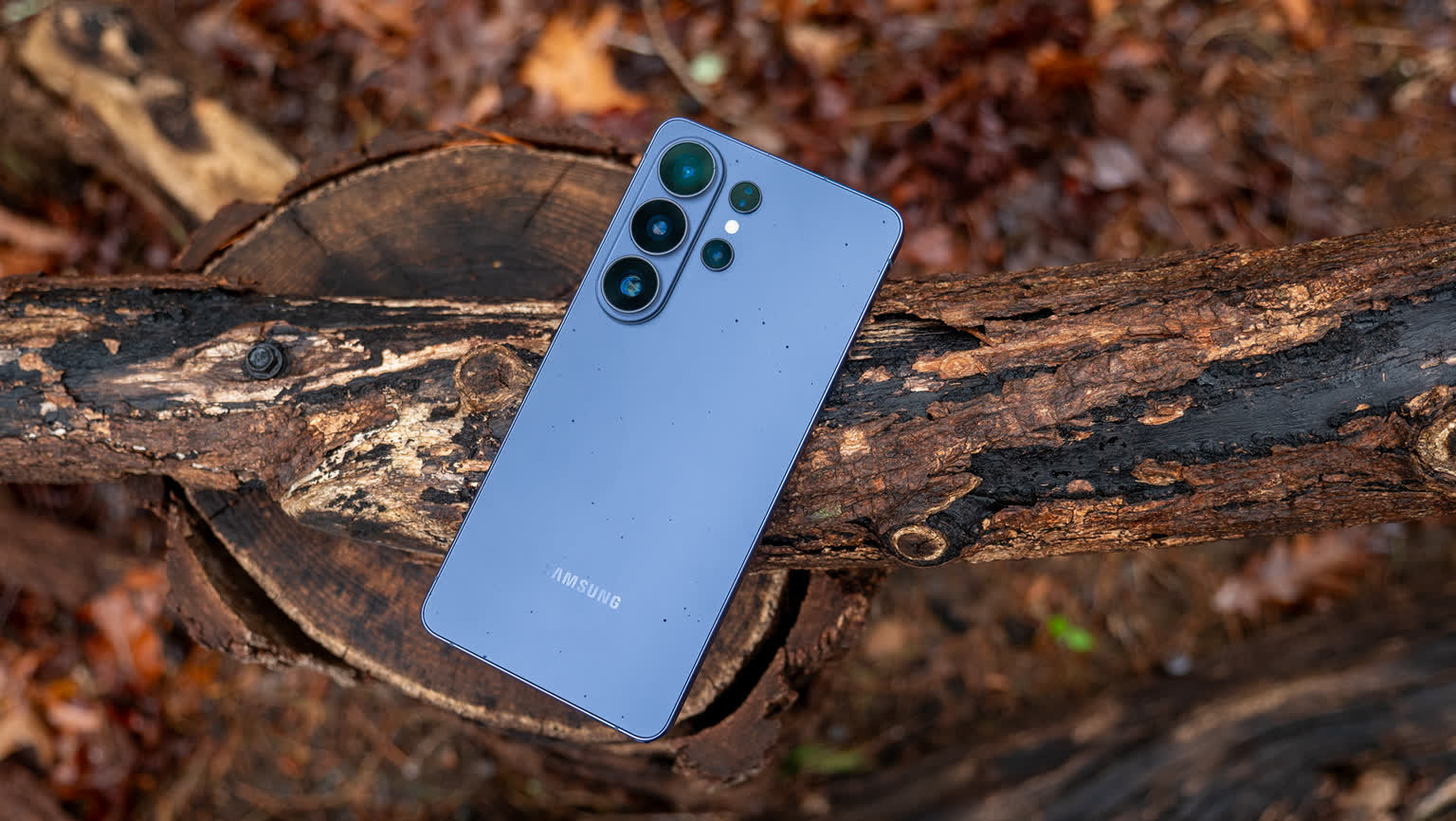 Blue Samsung smartphone resting on a rough log outdoors, camera module visible on the back.