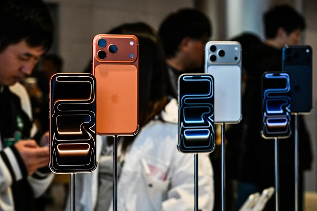 Multiple iPhones on display stands at a tech event, in orange, white, and blue colors with blurred crowd in the background