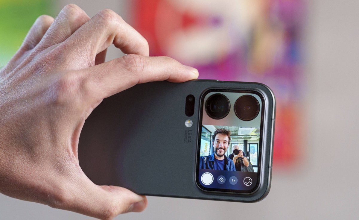 Hand holding a smartphone in a protective case; the rear display shows a person taking a selfie in the camera app.