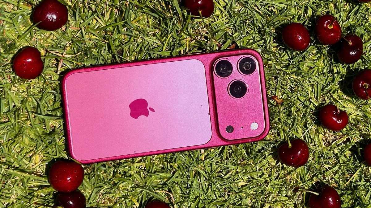Pink iPhone with triple-camera system lying on green grass among red cherries, top-down view, outdoor scene with bright lighting.