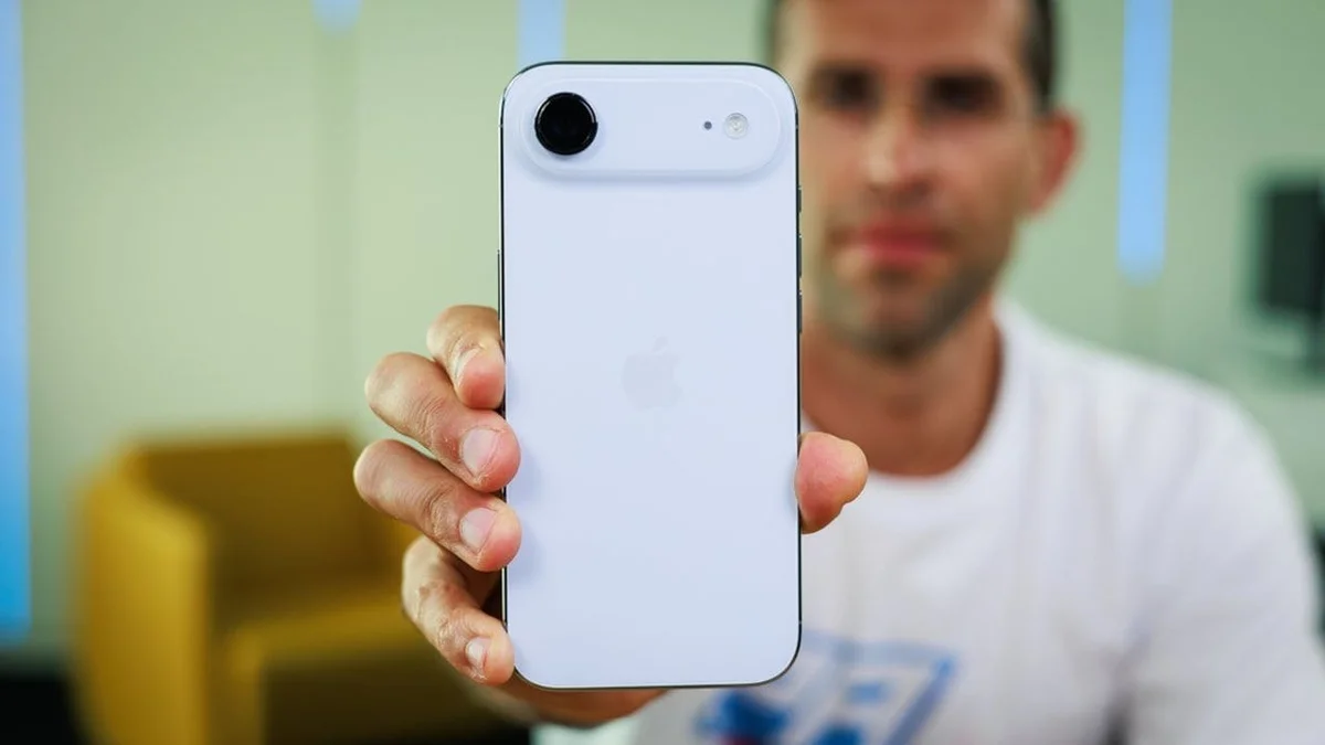 Person holding up a white iPhone with the back camera toward the viewer in a blurred indoor setting.