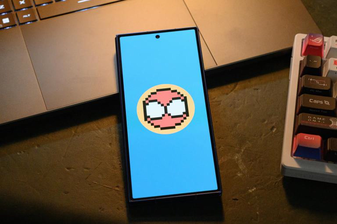 Phone on a desk showing a blue wallpaper with a pixel art circular emoji at center (pink face with white eyes).