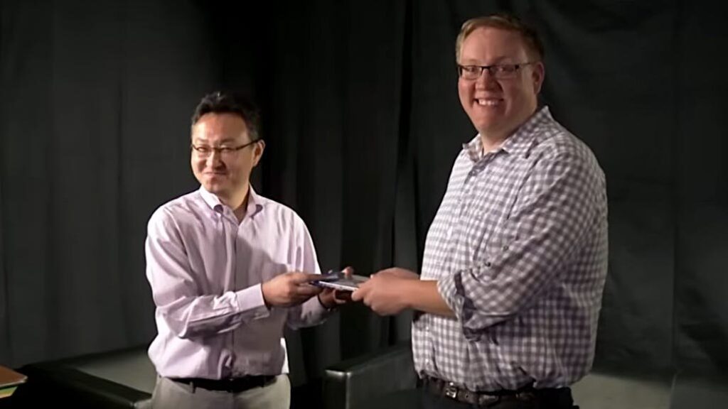 Two Men Stand on a Stage Exchanging a Small Plaque or Award Both Smiling As They Shake Hands