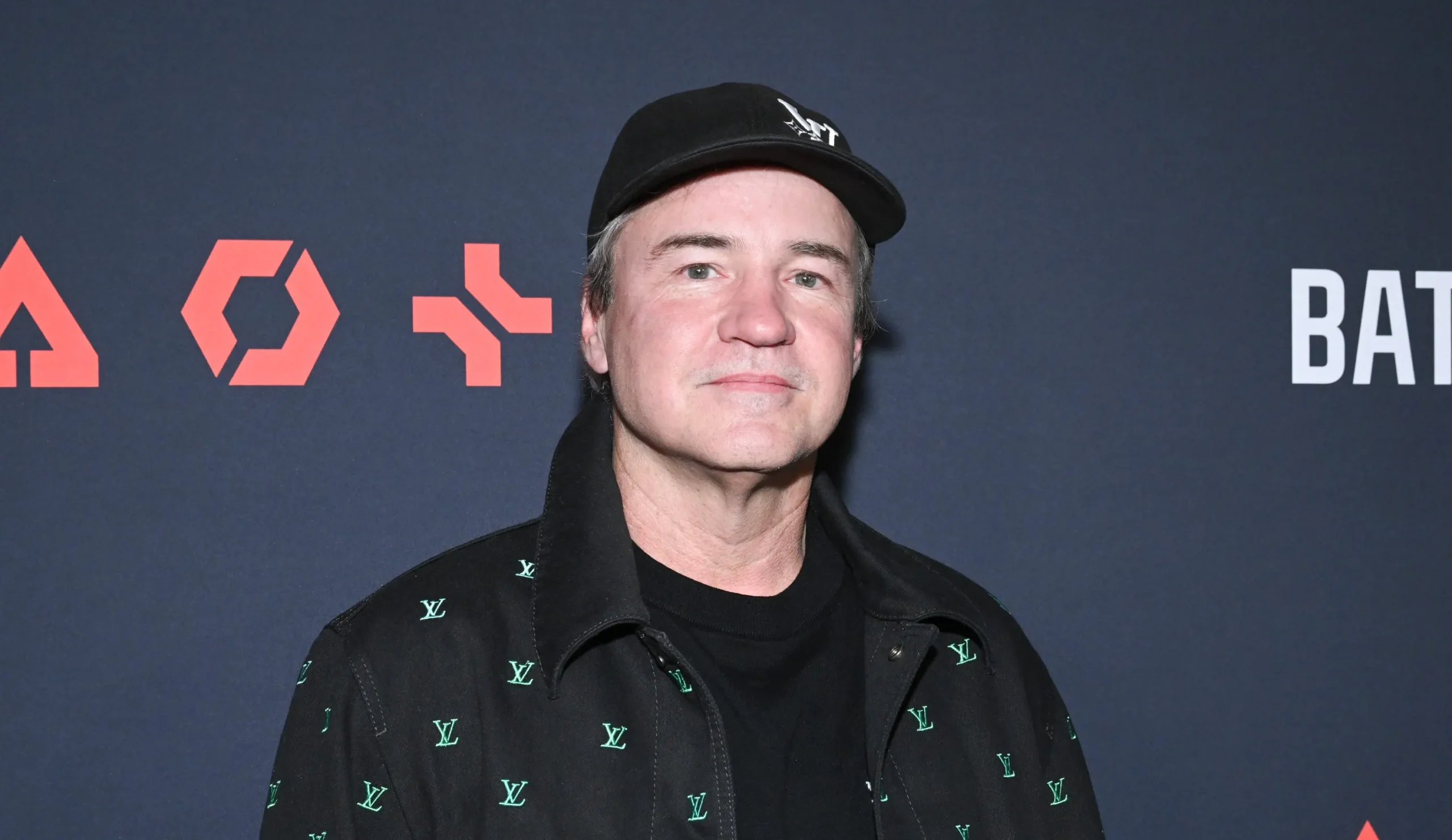 Man in a Black Cap and Black Jacket with Green Lv Logos Poses Against a Dark Blue Backdrop with Red Graphics