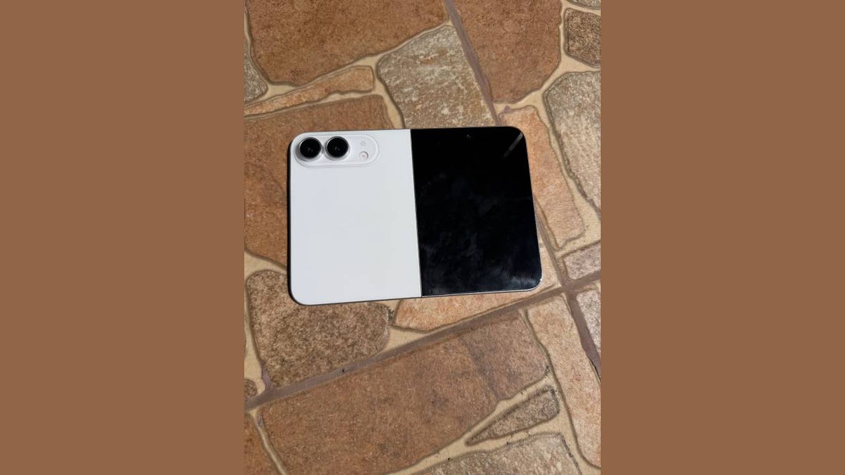 1000049413 Two-tone smartphone with a white left half and black right half resting on a stone-tile floor, showing a dual-camera module in the corner.