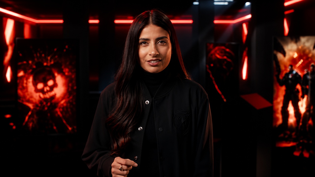 Woman with Long Dark Hair in a Black Varsity Jacket Stands in a Dark Room with Red Neon Lights and Posters in the Background As She Faces the Camera