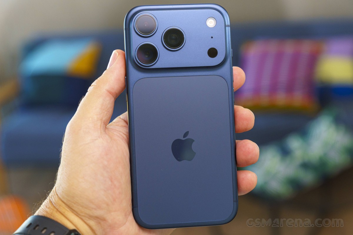 1000049382 Blue iPhone held in a left hand, showing the triple-camera setup on the back and Apple logo.