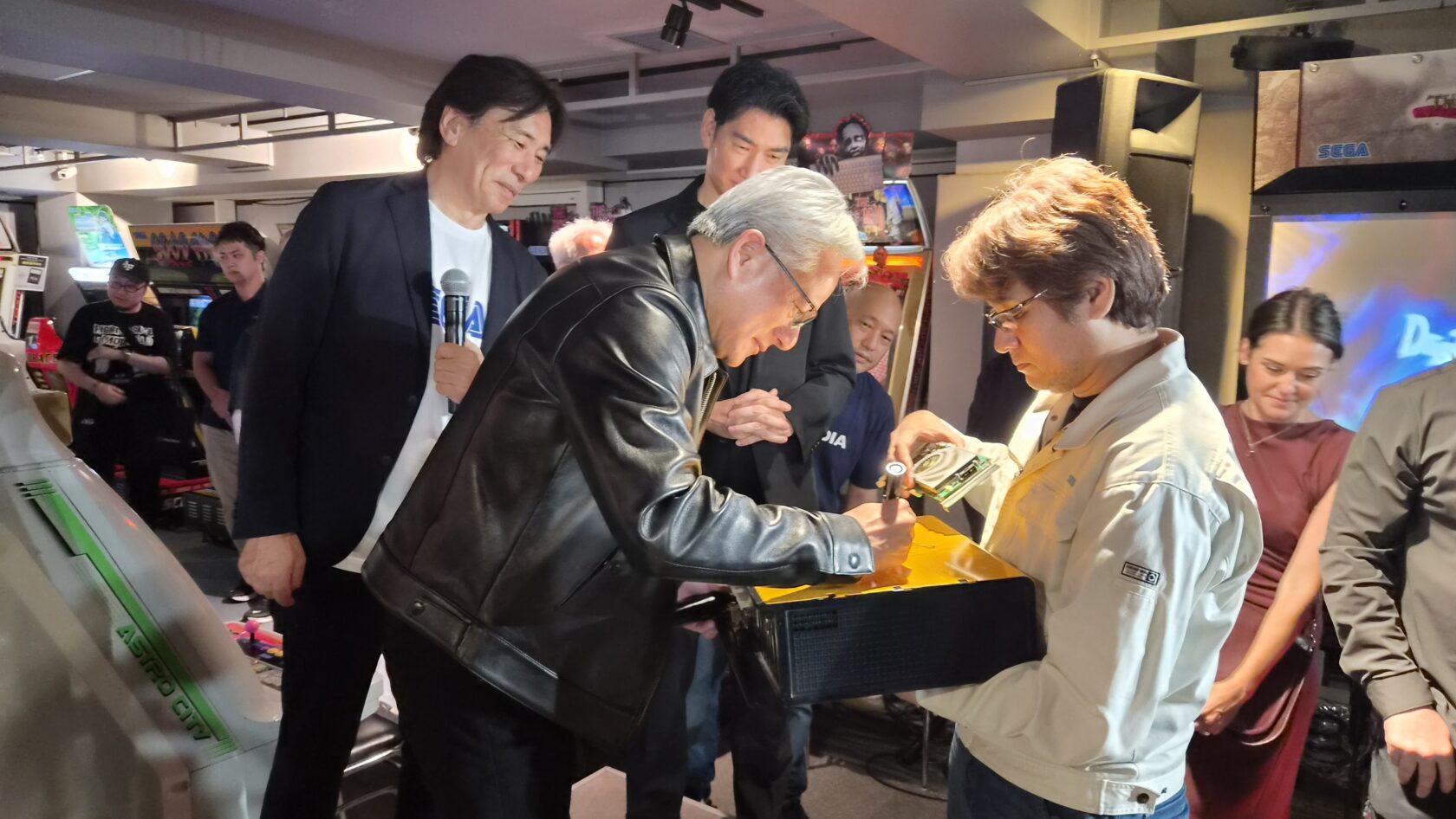 De3db269 98cd 4541 a47e 3bc4c91a4420 1680x946 1 Man in a Black Leather Jacket Signs an Item for a Person Holding It While a Crowd Watches at an Arcade Event with Arcade Machines in the Background