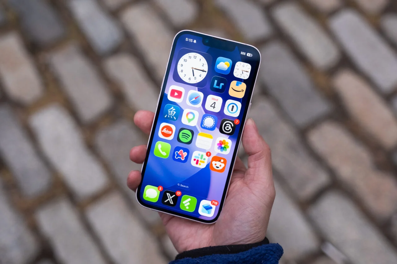 Hand holding a smartphone showing a colorful app grid on the home screen over a cobblestone background.