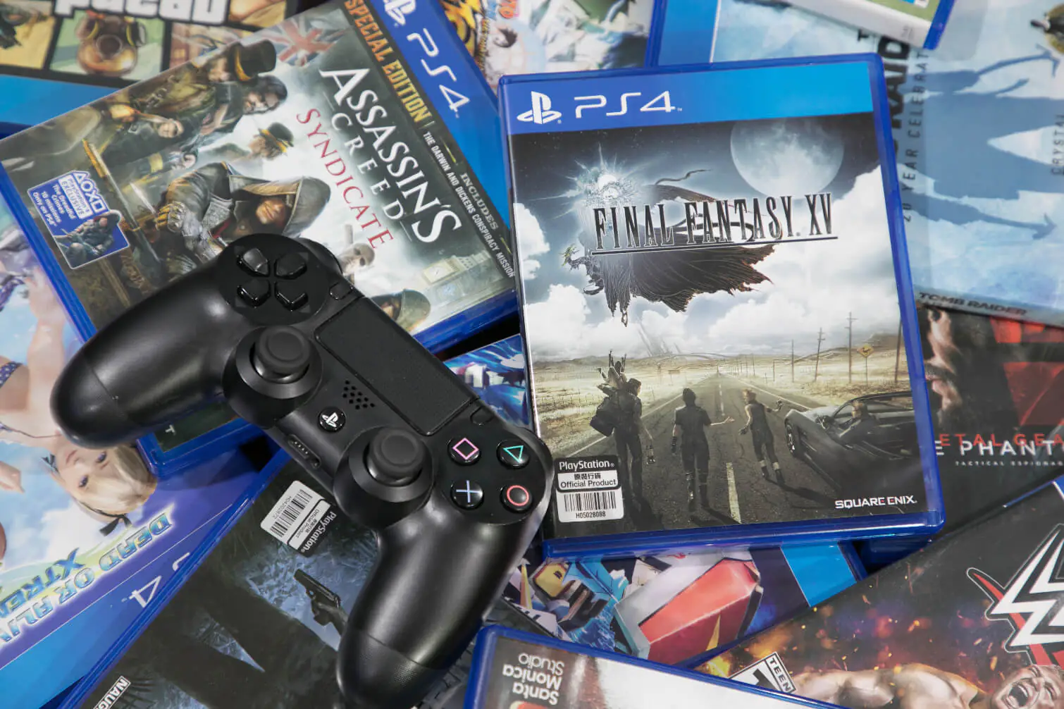 Black Ps4 Controller Resting on a Pile of Blue Ps4 Game Cases Including Final Fantasy Xv and Assassin's Creed Syndicate