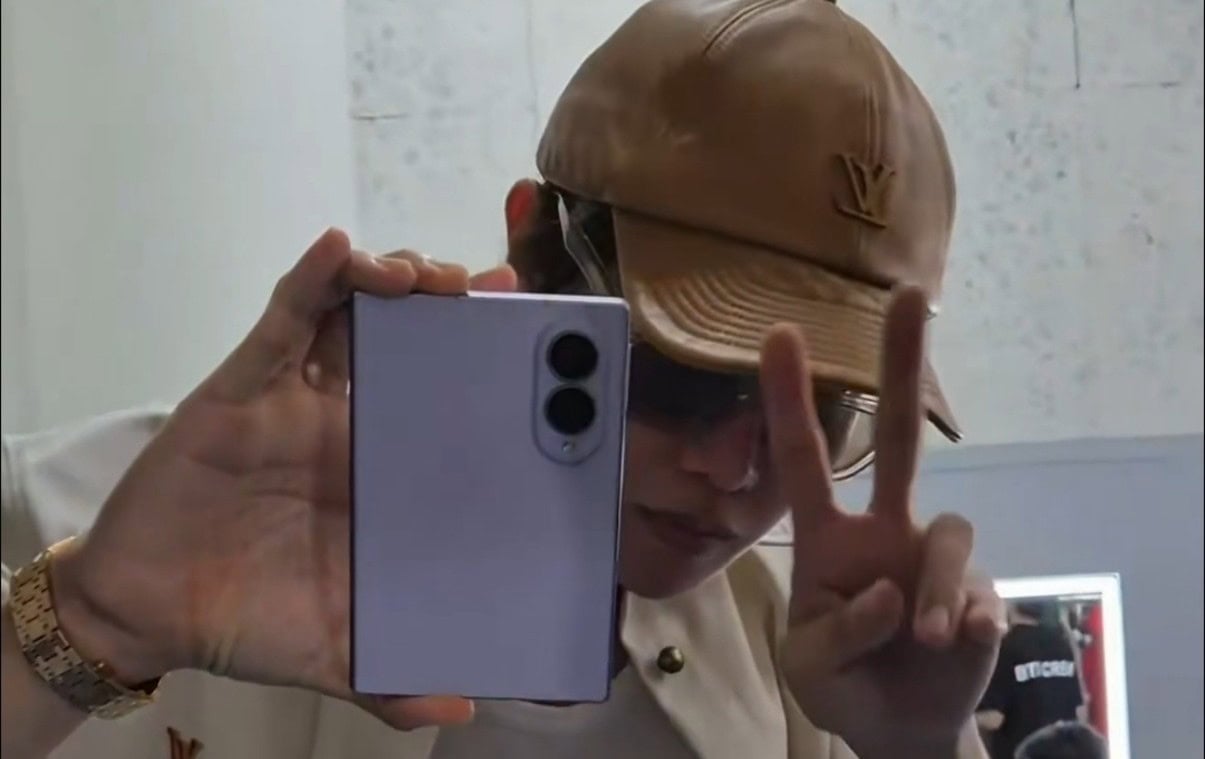 Person takes a mirror selfie with a blue smartphone, wearing a tan cap and glasses, flashing a peace sign with two fingers.