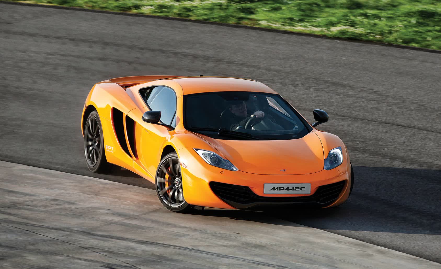 1000042758 Orange McLaren MP4-12C sports car speeding on a race track, turning around a curve with green scenery in the background