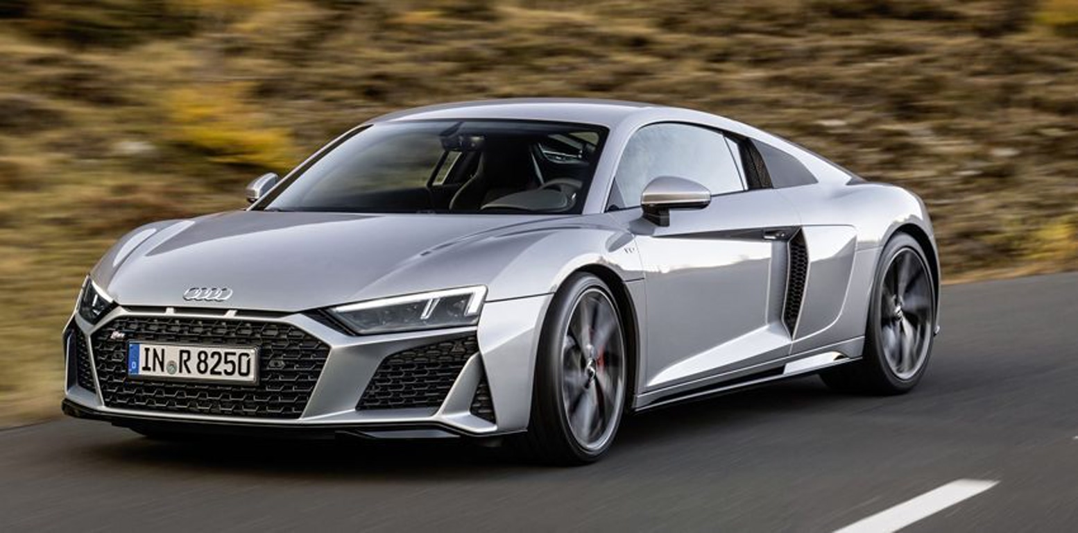1000042760 Silver Audi R8 sports car driving on a highway with a blurred background to convey speed and motion, front three-quarter view.