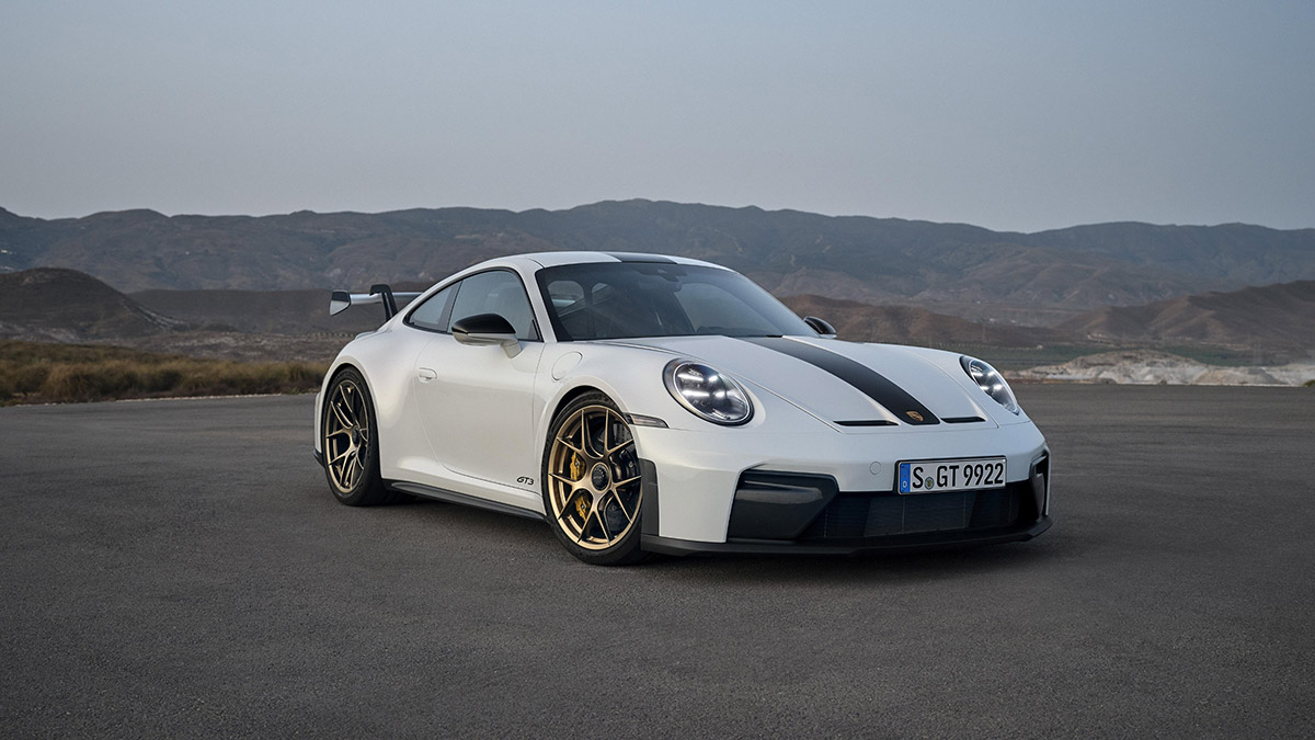 1000042745 White Porsche 911 GT3 with a black racing stripe and gold wheels parked on a flat runway with desert mountains in the background