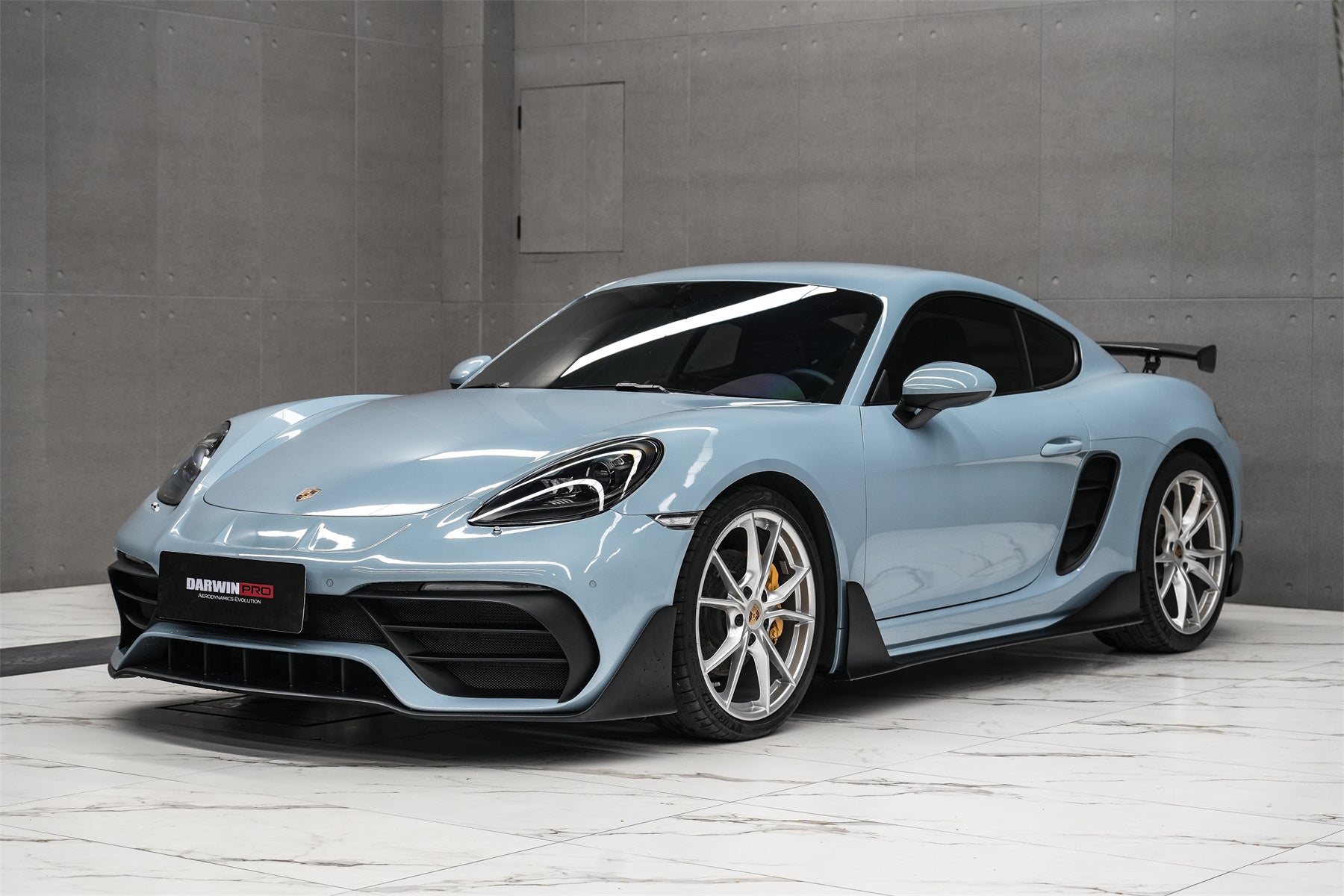1000042750 Pale-blue Porsche sports car with large rear wing, shown in a minimalist concrete showroom on a marble floor.