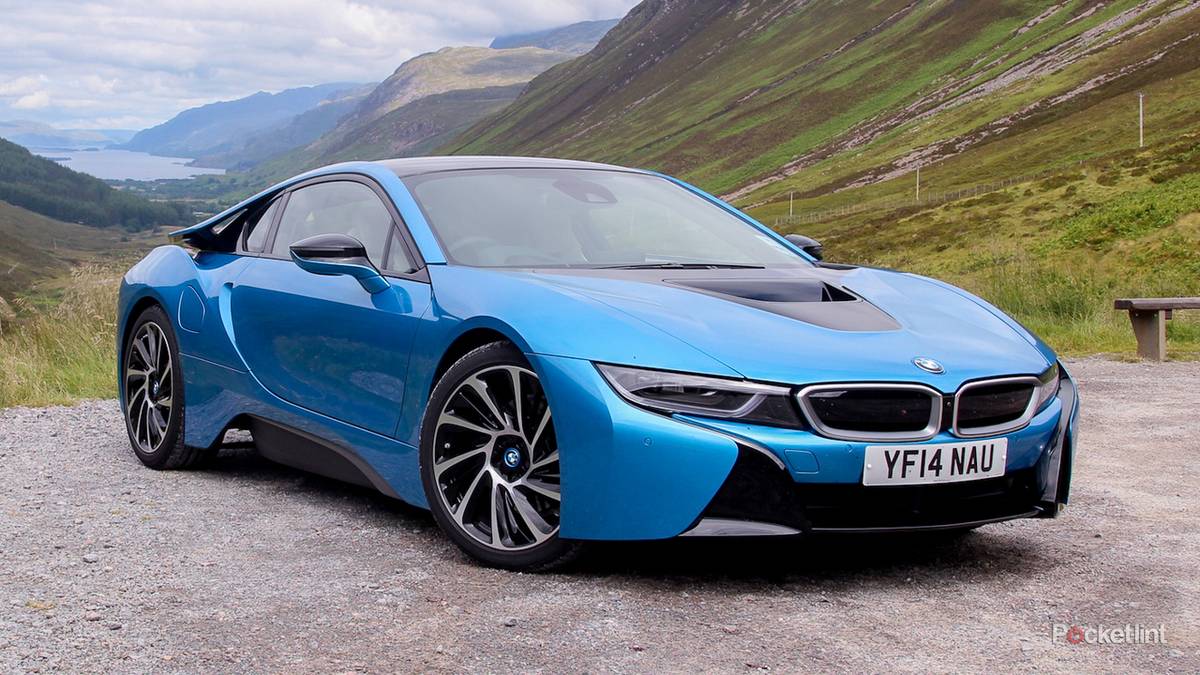 1000042755 Blue BMW i8 sports car parked on a gravel overlook with green hills and mountains in the background while a lake is visible in the distance.