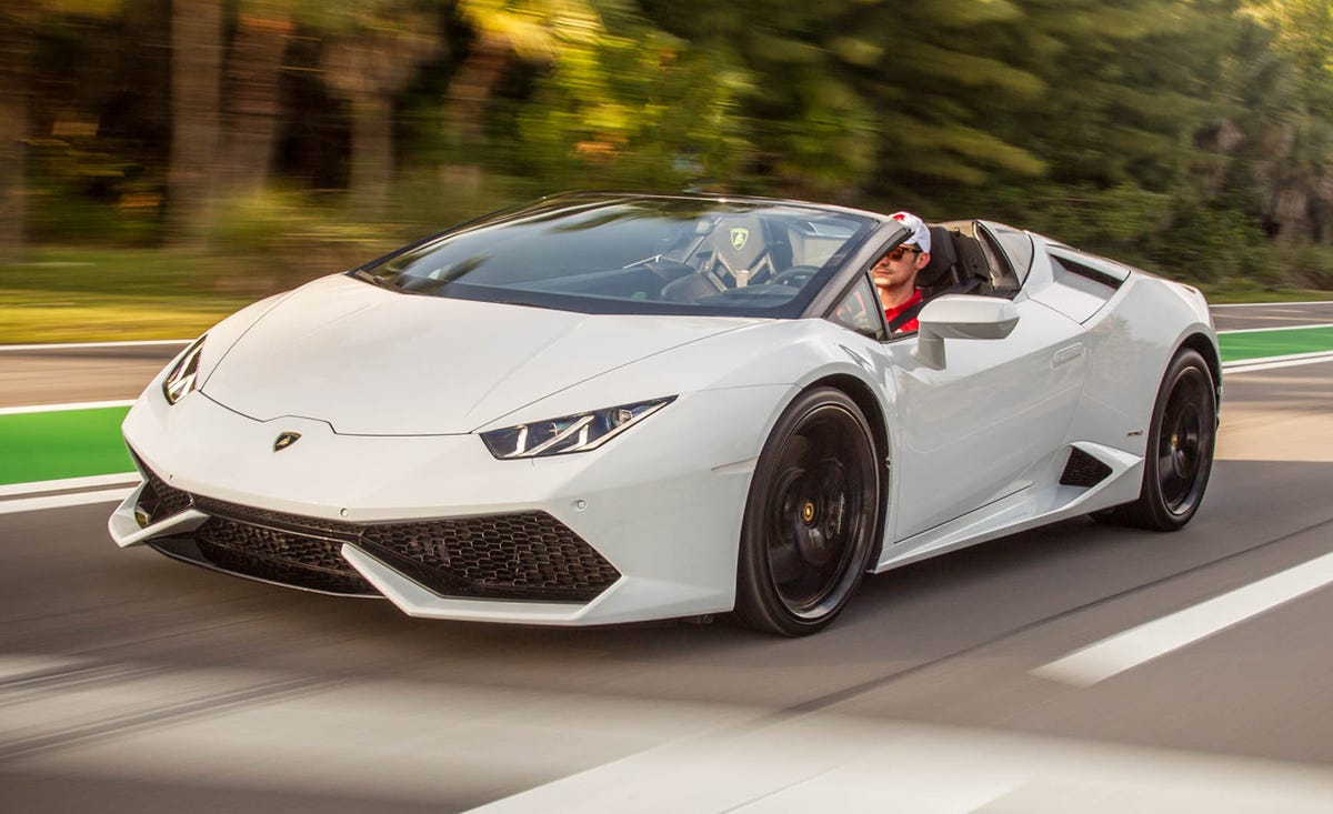 1000042748 White Lamborghini Huracán Spyder driving at speed on a road with a male driver in a red shirt and cap, convertible top down.